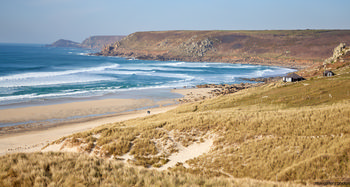 Sennen cove from dunes This landscape photograph captures Sennen Cove from the dunes on the coast of Cornwall, United Kingdom, in the afternoon during the early spring season. The image features rolling waves approaching the sandy beach, with the rugged hills of the Cornish coastline extending into the distance. The natural beauty of Sennen Cove is highlighted by the presence of grassy dunes in the foreground and a few traditional cottages nestled near the shore. The scene showcases the untouched nature of the area, with the beach and shoreline characterizing the unique coastal environment of Cornwall.